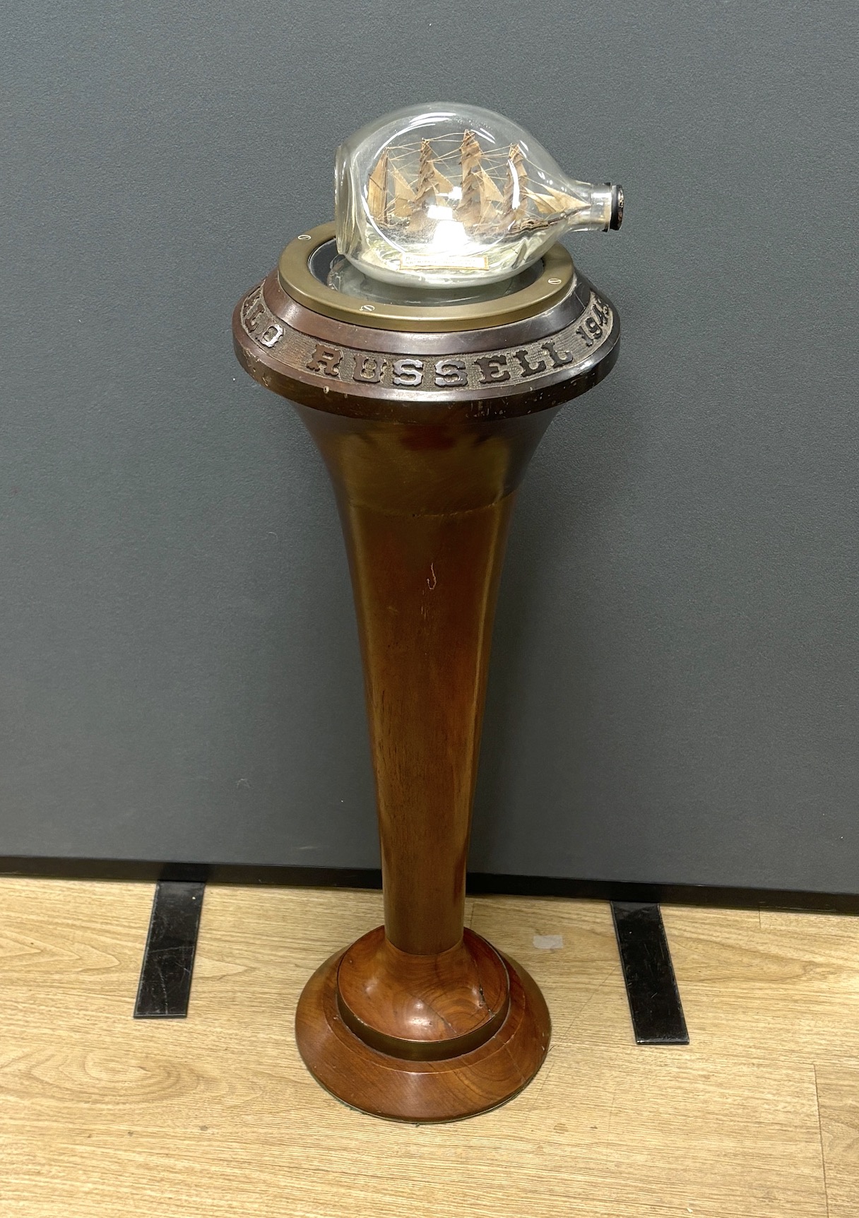 A ship’s compass in a gimbal, believed to be from the tall ship The Archibald Russell, mounted on a floor standing mahogany stand with carved attribution around the edge, together with a ship in a bottle of the barque in